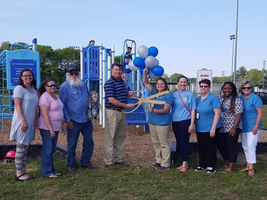A group of adults stands at a playground holding oversized scissors and balloons during a ribbon-cutting event.