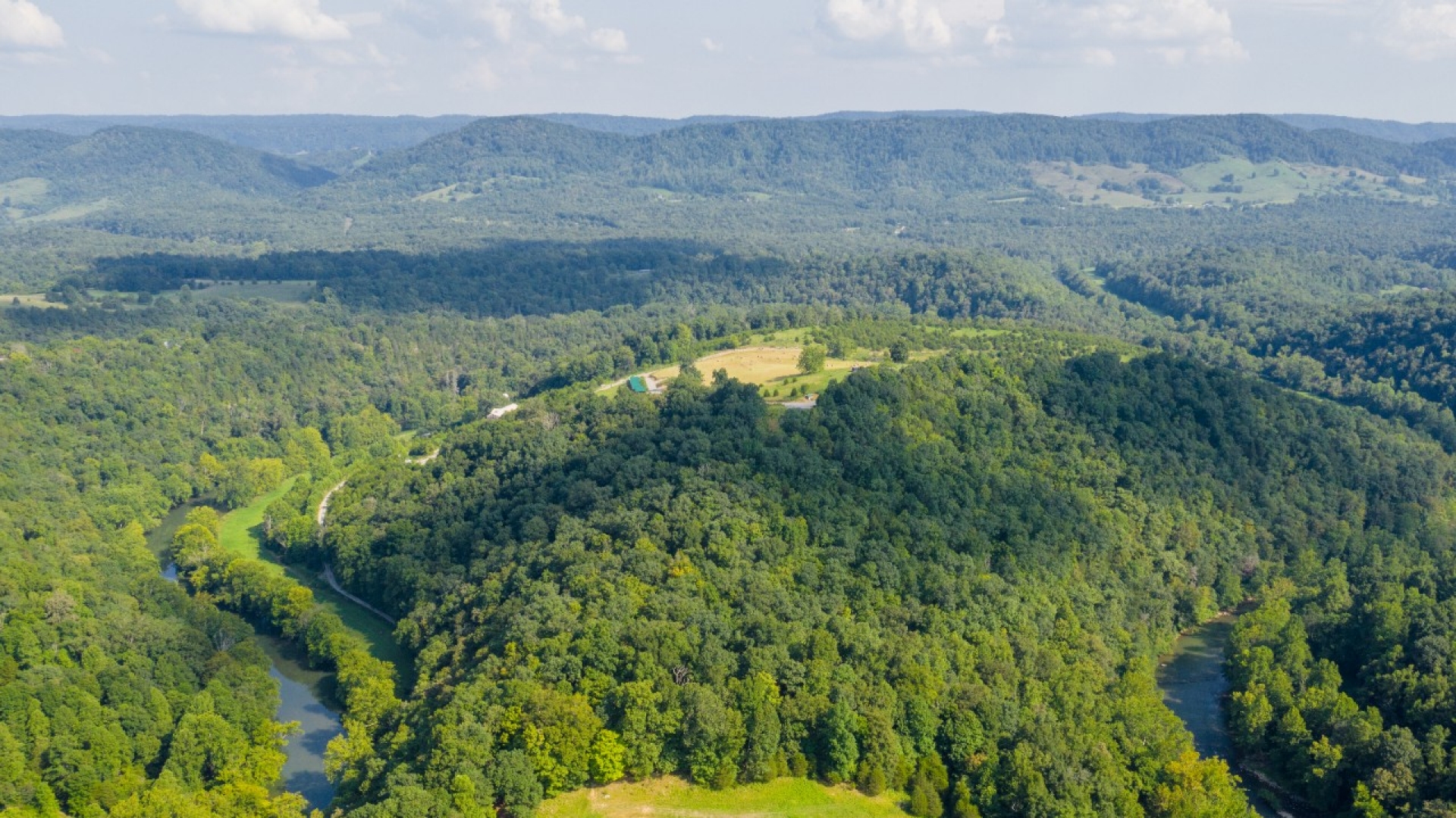 An aerial view shows forested hills and a winding river cutting through a green rural landscape.