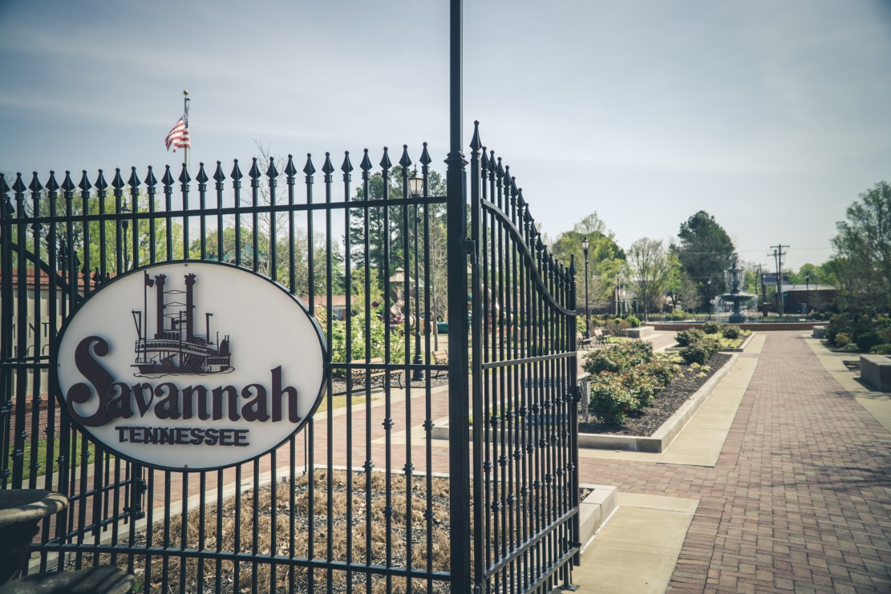 A gated entrance opens to a landscaped town park with brick walkways, gardens, and a central fountain.