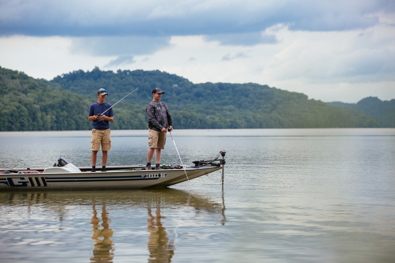 Two people fish from a small boat on a calm lake, with forested hills and cloudy skies in the background.