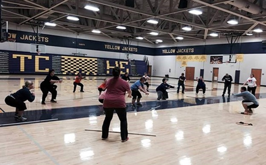 A group of people participates in a fitness class inside a school gymnasium, spread out across the basketball court.