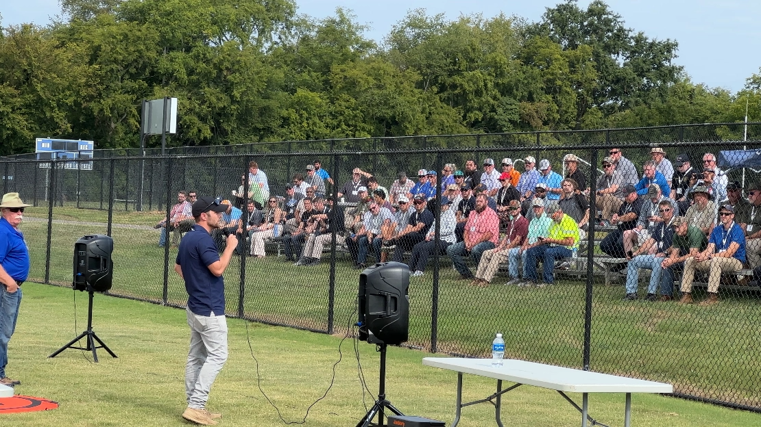 Speaker addressing an outdoor audience seated behind a fence at a drone event, with audio equipment and a table set up on the field.