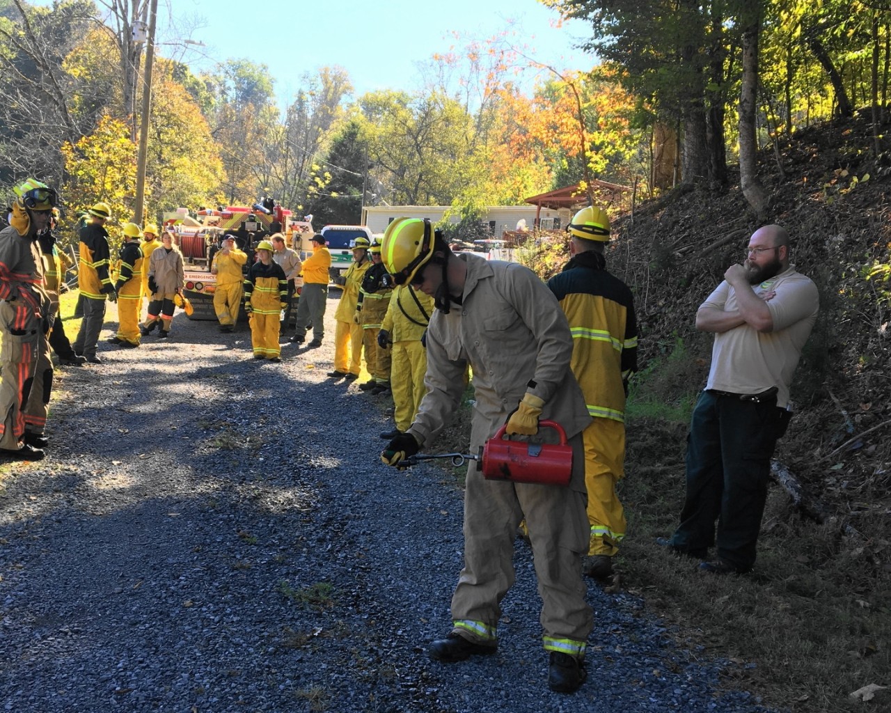 Firefighters in yellow and beige gear stand on a gravel path near a red emergency vehicle in a forested, autumn setting.