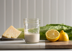 Homemade Caesar dressing in a small jar on a table with a lemon, romaine lettuce and parmesan cheese next to it
