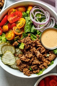 a bowl with hamburger meat, lettuce, onions, pickles, tomatoes and a side of sauce