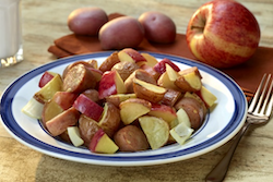 Roasted potatoes, apples and sausage in a bowl; in the background are whole potatoes and an apple