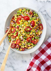 corn and edamame succotash salad in a large bowl with wooden utensils on a marble counter with a gingham towel next to it