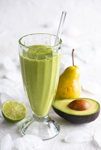 a green smoothie in a glass with a glass straw and a fresh pear, an avocado and half a lime on the counter