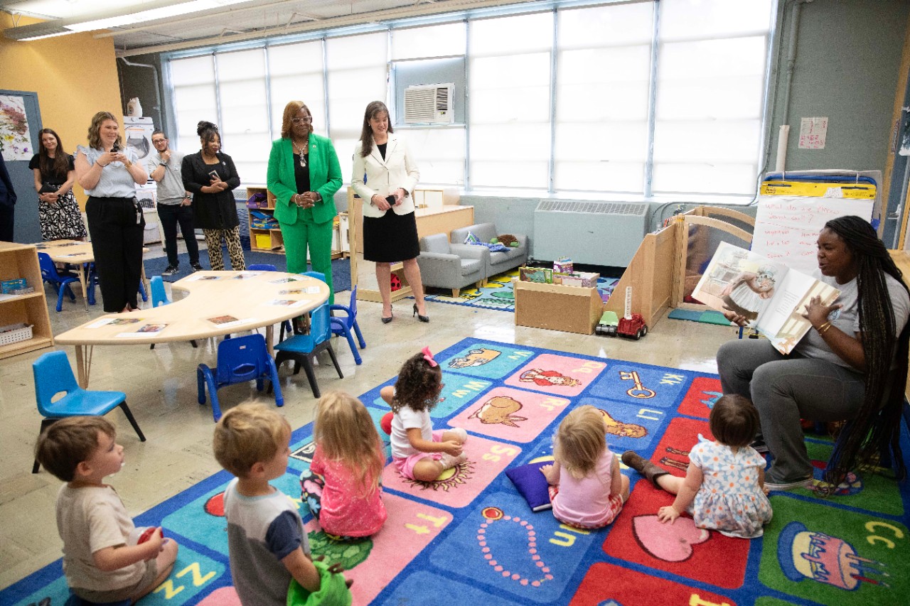 Deniece Thomas and Candice McQueen Teacher reads a storybook to a group of young kids on a mat as several adults stand and watch