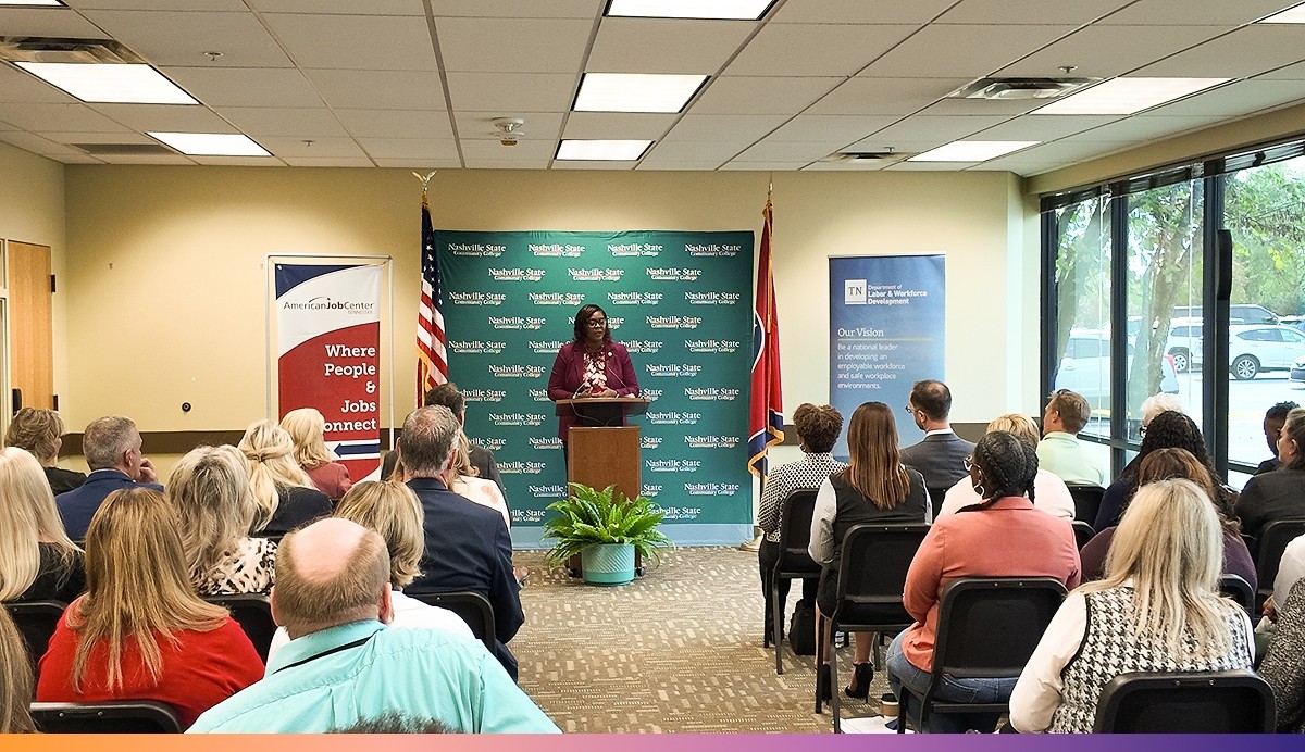Woman speaks at podium in front of an audience. Behind her is signage for the American Job Center, Nashville State Community College, and the Tennessee Department of Labor and Workforce Development