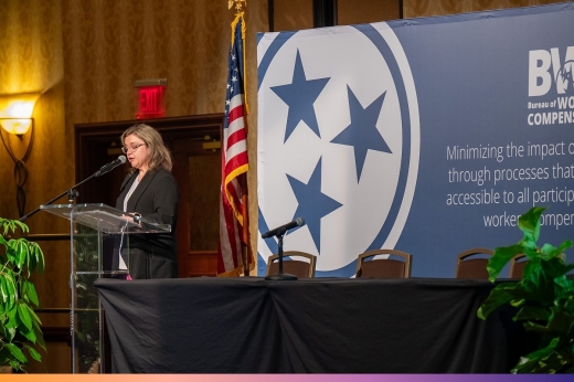 Woman speaks at a lectern in front of a large Bureau of Workers' Compensation banner