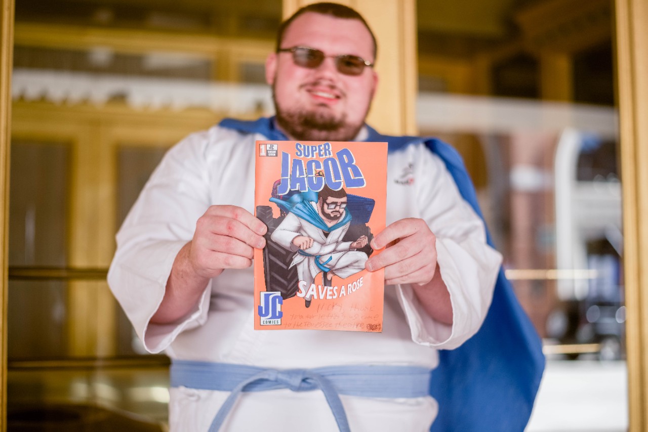 JacobCollins Jacob is holding out his comic book Super Jacob. The comic book has an image of a superhero wearing a white costume with a blue belt and cape