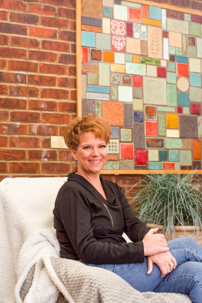 DSC_7871 Juli, a 51 yo woman with short red hair, sits in front of a colorful mosaic painting outdoors