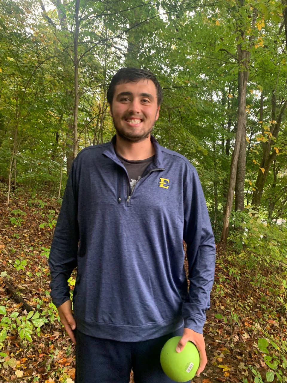 Outdoors Nick A medium complected man in an ETSU polo smiling outside and holding a green ball