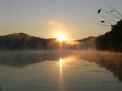 The sun sits at the crest of hill surrounding a foggy lake