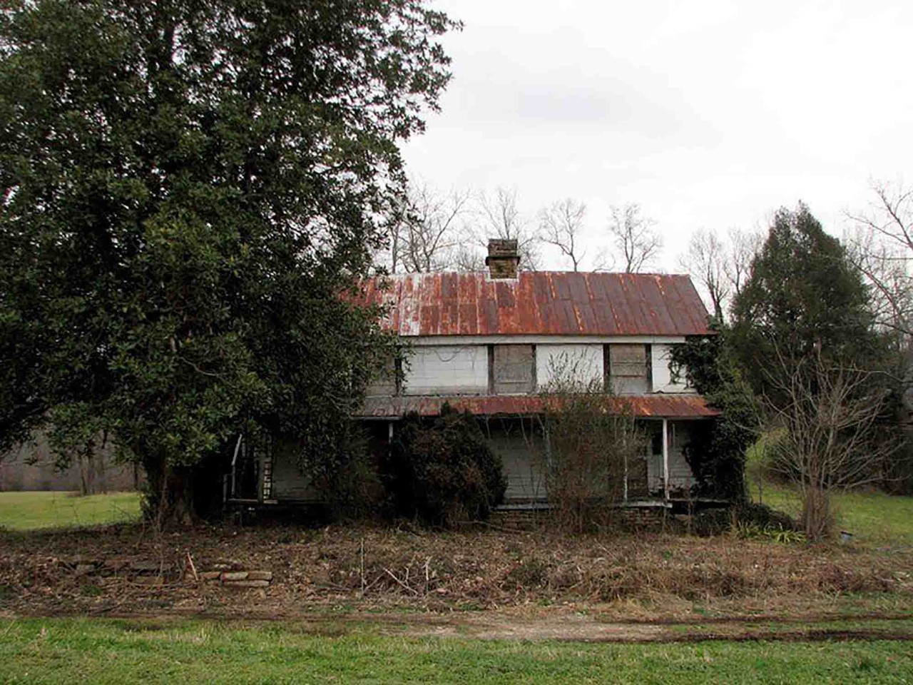 The Stonecipher Kelly House when purchased in 2014 for restoration. Image of The Stonecipher Kelly House when purchased in 2014 for restoration. Image shows an old run-down house with a rusted roof and front porch. The lawn is brown, and the front of the house is overgrown with vegetation.
