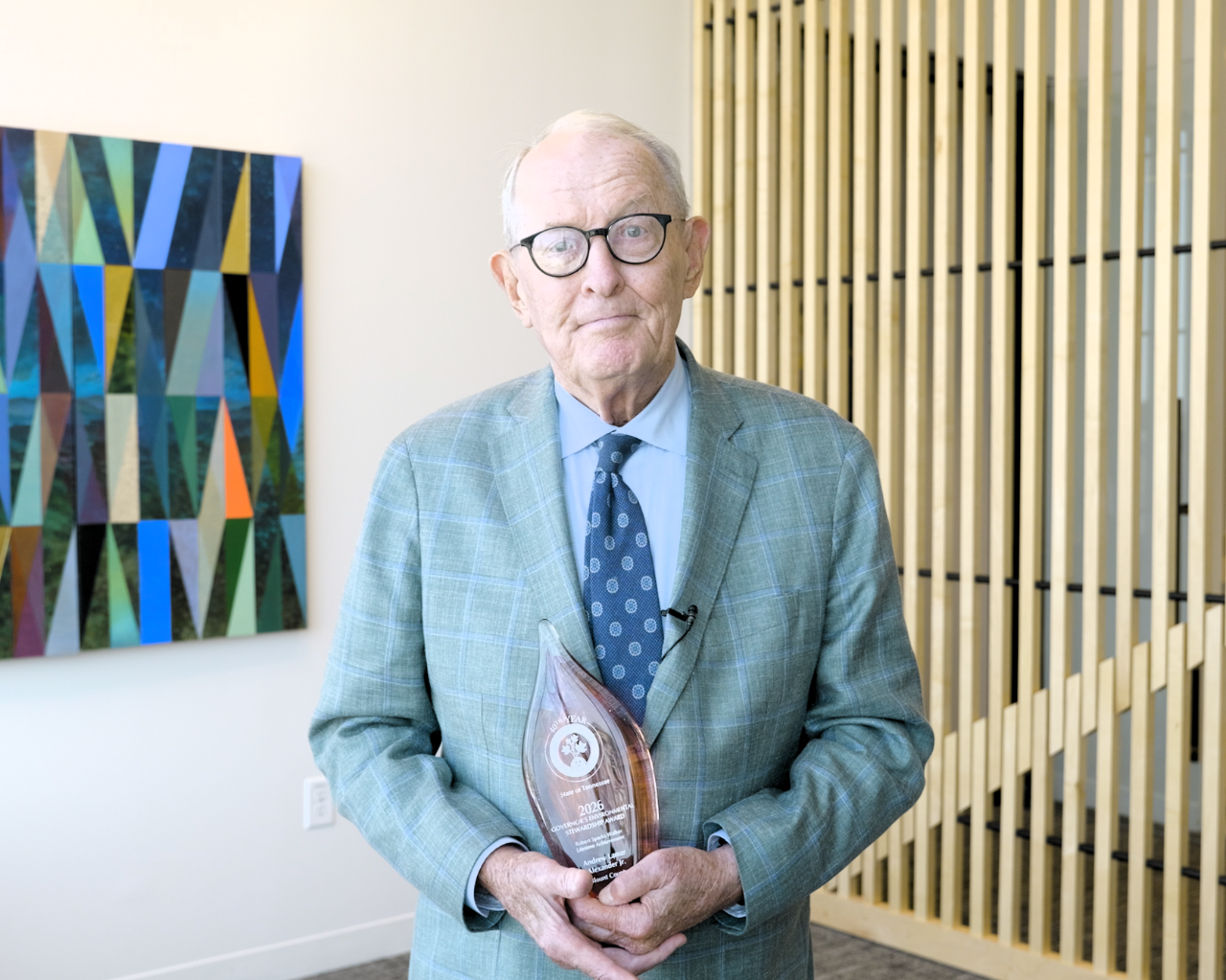 tdec-news_lamar-alexander-gesa-award A man standing indoors, wearing a light-colored plaid suit jacket, and glasses, holding a clear, teardrop-shaped award that reads "2026 Governor's Environmental Stewardship Award, Robert Sparks Walker Lifetime Achievement, Andrew Lamar Alexander, Jr., Blount County." Behind the individual is a geometric multicolored artwork on the wall and a vertical wooden slat divider.