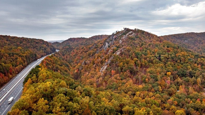 tdec-news_devils_racetrack_chuck_sutherland An aerial view of a highway curving through a dense forest of autumn foliage, with shades of green, yellow, orange, and red leaves covering rolling hills. A rocky ridgeline rises prominently near the center of the image under a cloudy sky.