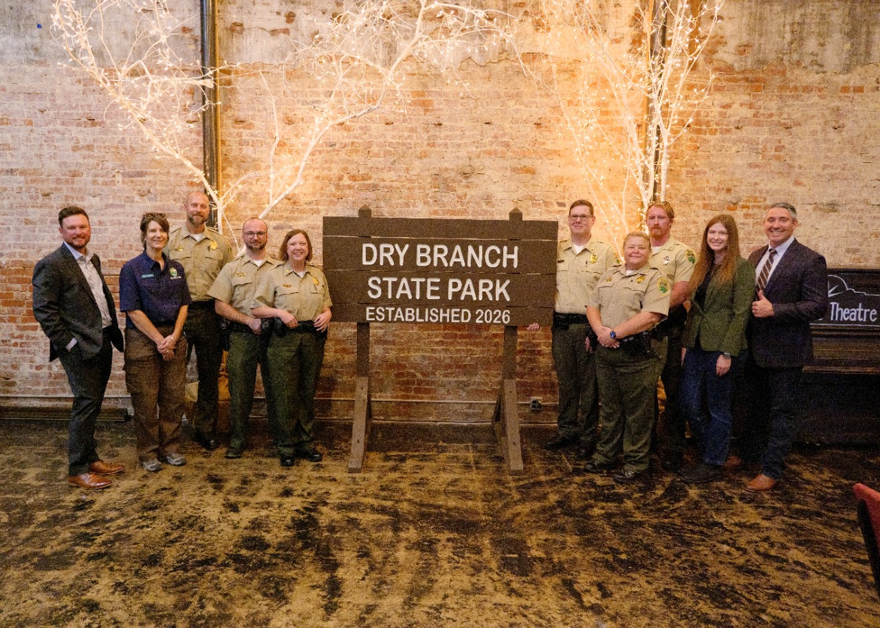 tdec-news_dry_branch_sp_dedication Group of people, including Tennessee State Parks rangers in uniform, standing on either side of a large wooden sign that reads "Dry Branch State Park Established 2026." The group is posed in front of a brick wall decorated with illuminated tree‑branch lights.