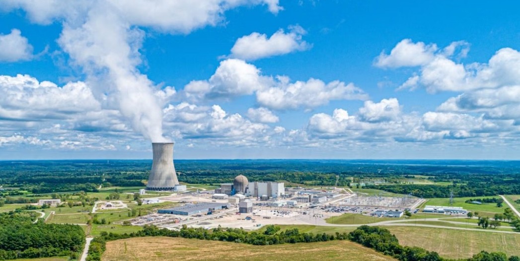 Aerial view of a commercial nuclear power plant, showing a cooling tower emitting water vapor, reactor buildings, and supporting infrastructure surrounded by farmland and forest. The image illustrates Tennessee's nuclear energy infrastructure.