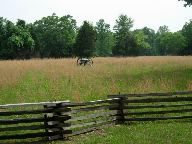 A Civil War cannon sits in the middle of a grassy field bordered by a split rail fence