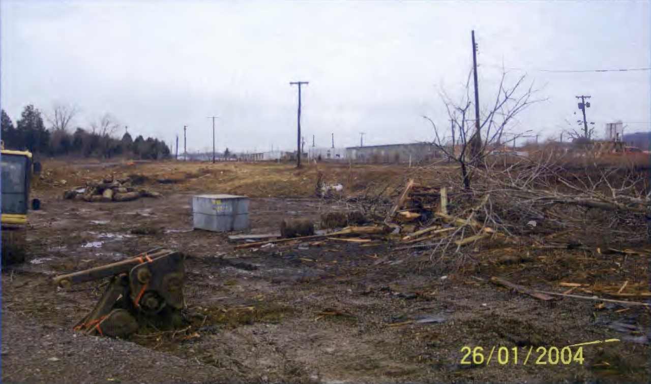 A B-25 box containing radioactive material on the Atomic City Auto Parts site. A B-25 box containing radioactive material removed from the Atomic City site sits in a dirt plane along with rusty equipment, dead trees, piles of dirt and cylinders, and construction supplies.