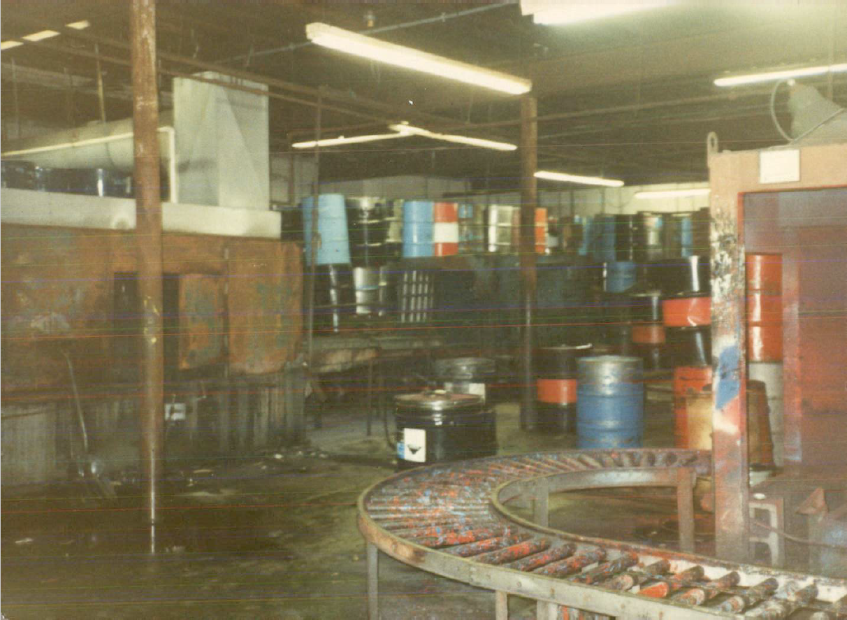 Dixie Barrel and Drum facility before demolition. An old photo of the paint-stained conveyor belt inside the Dixie Barrel & Drum facility. Cleaned and restored drums sit on shelves and the floor in the background, and puddles on the concrete slab floor reflect rusty iron beams supporting the building.