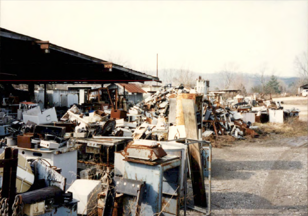 Joyner Scrap Yard contents before radiological contamination removal. Joyner scrap yard including heaps of miscellany on the bare ground. Items include rusted metal parts, large boxes, and indistinguishable materials.