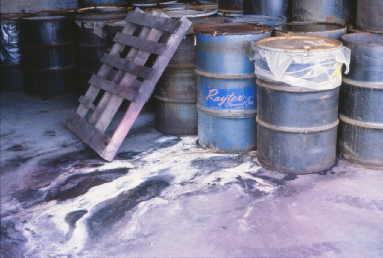Leaking drums in the former Morningside Chemical facility Leaking drums in the Morningside Chemical facility. A wooden pallet is leaned against a dozen drums, mostly gray and rusted with a blue drum labelled "Raytex Chemical" in the foreground that appears to be leaking a white oxide material all over the concrete floor.