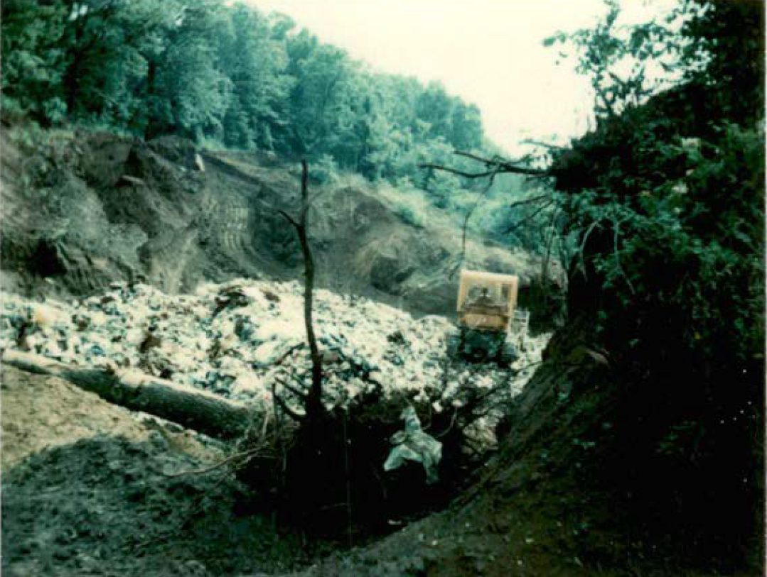 Exposed contents of the municipal and industrial Red Ridge landfill before cap was installed. Trash and miscellany fill an open pit in the ground. An uprooted tree stump sits on its side in the foreground with trash embedded in the dirt-caked roots. A tractor plows over the trash pile in the background.