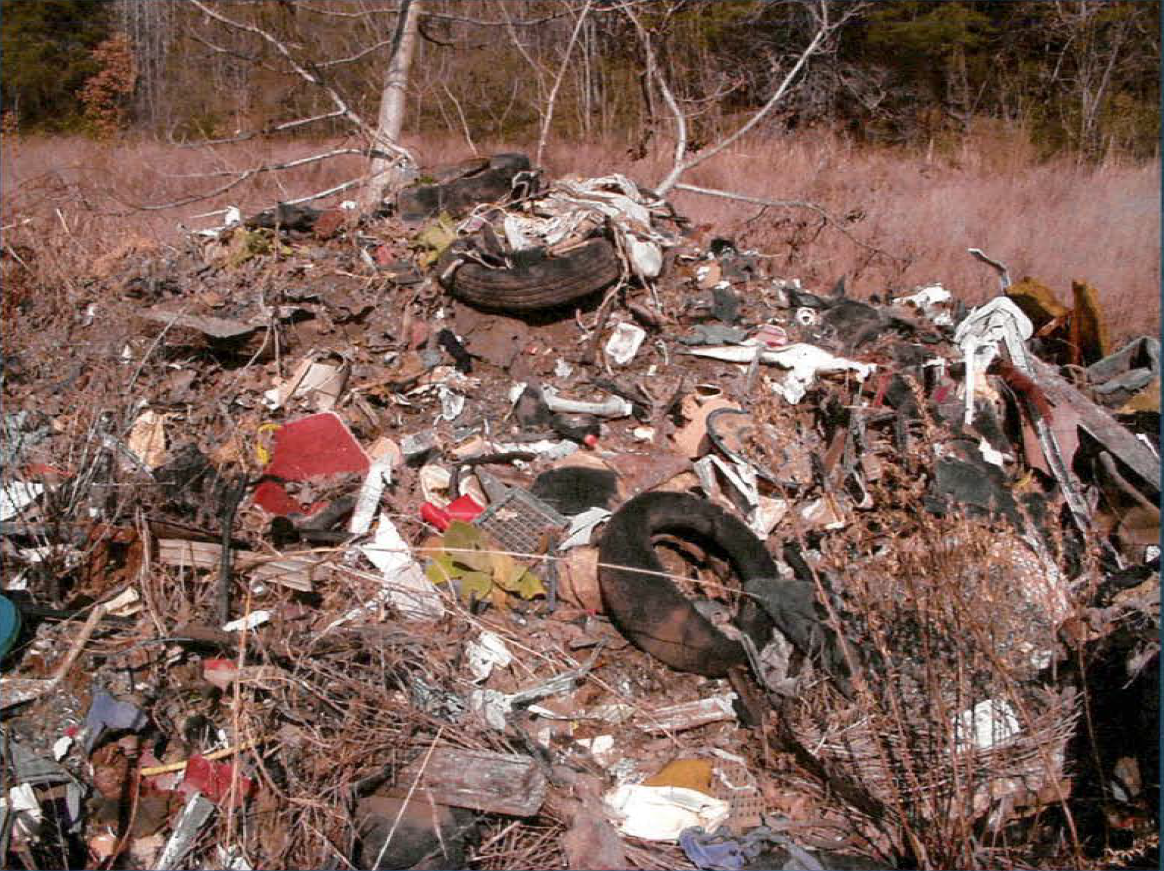 Rockwood Iron & Metals automobile shredding residue debris pile. An uncovered debris pile of automobile shredding residue containing dirt, tires, shards of metal, and seat fluff; surrounded by trees and grass.