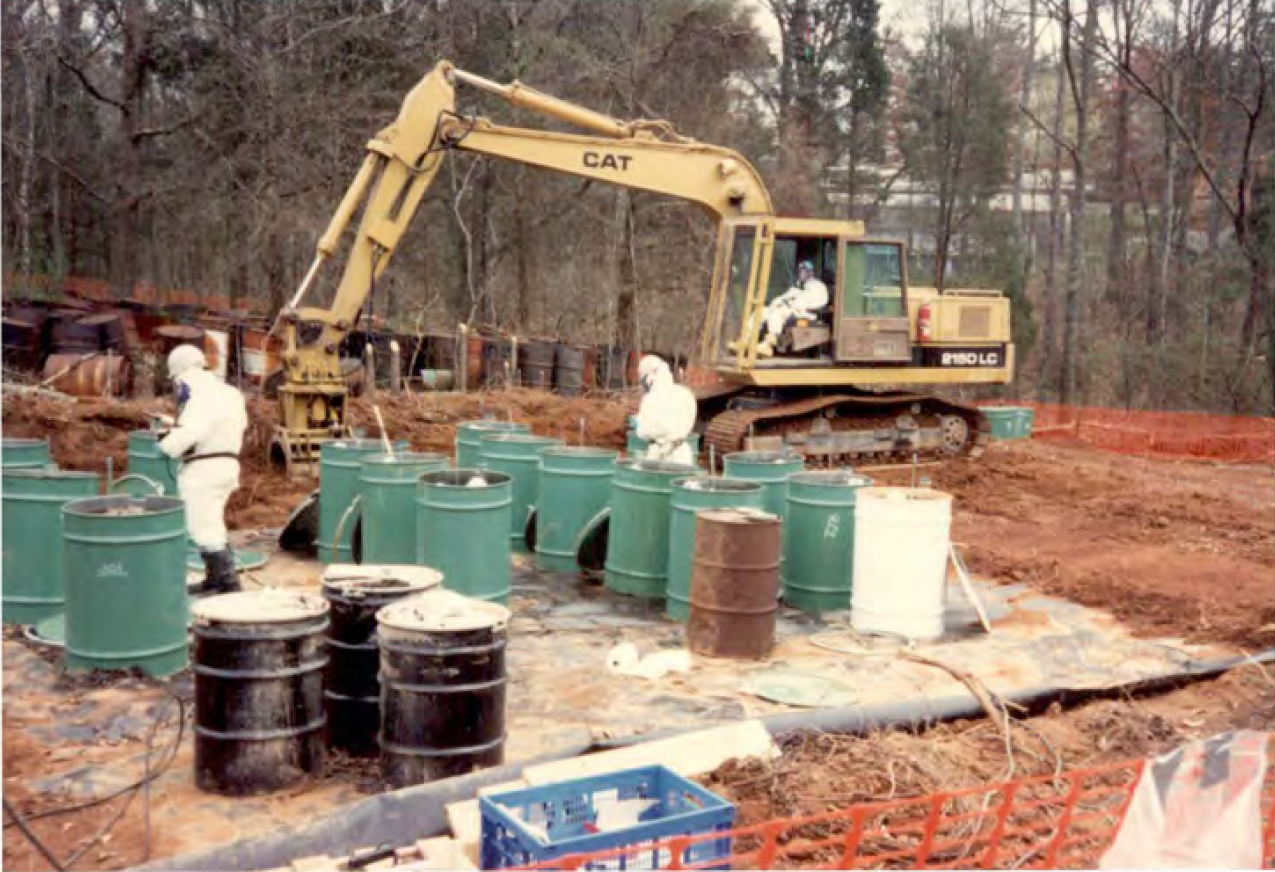 EPA cleanup of hazardous waste in drums stored at the Roscoe Fields site. Cleanup of dozens of green and brown steel drums is conducted by workers in white haz-mat suits.
