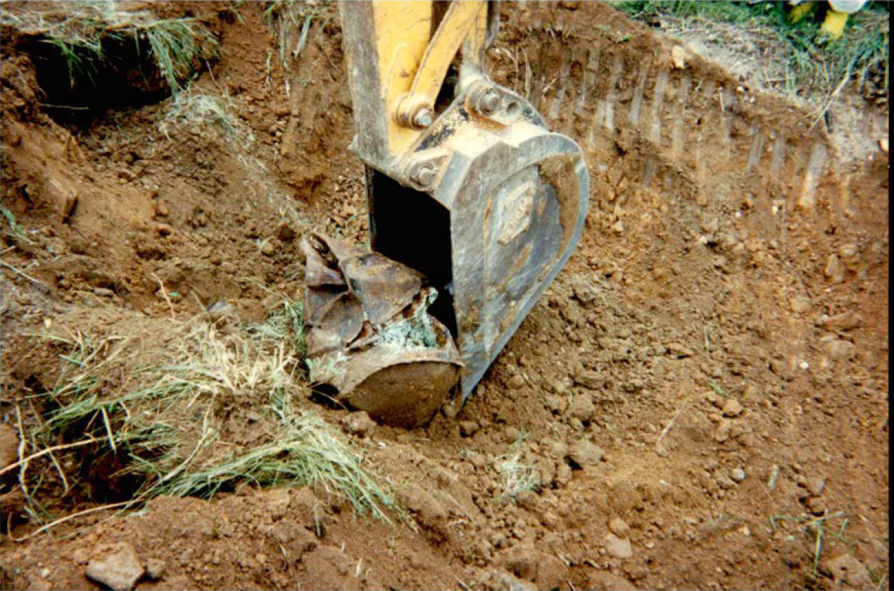 Excavator digs out a crushed drum containing unknown materials on the Wall Tube & Metals site. An excavator bulldozer rests the digging arm on a crushed rusty metal drum containing a strange greenish oxide in a large dirt pit.