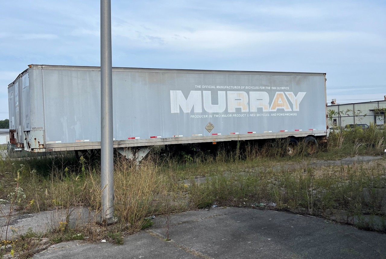 Murray Ohio dilapidated shipping container at the facility. A grassy parking lot with an old truck with faded and bleached decal reading "The official manufacturer of bicycles for the 1984 Olympics: Murray: producer in two major product lines: bicycles and powermowers."
