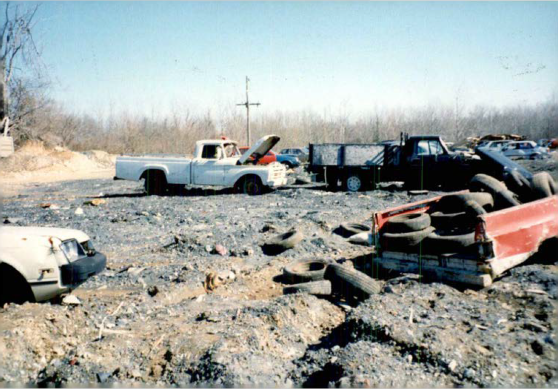 fiberfine auto yard Auto salvage yard on the Fiberfine site. A gray, lumpy, topographically uneven gravel yard hosts sagging cars. A white 1980s Chevy truck in the background with its hood parked. Old tires spill out of a red truckbed in the foreground.