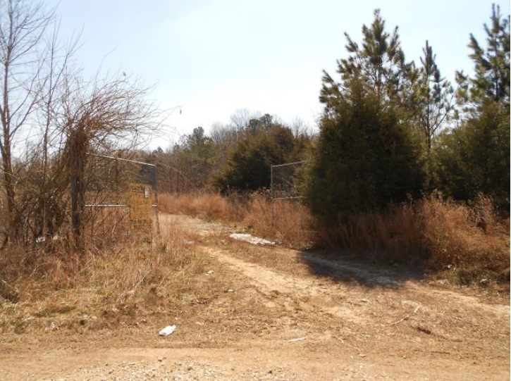 Henry County Boneyard front gates Dirt road entryway to the Henry County Boneyard site featuring pine trees, dead grass, and an open chain link gate with yellow "no tresspassing" sign.