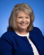 Tammy NEW headshot A woman with light brownish blond hair smiling against a blue background. She is wearing a white shirt with a blue knit textured jacket.