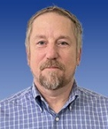 Mattox, Christopher_00639738_BA020003 A man with a slight smile set against a blue background. He has graying hair, a moustache, and a beard. He is wearing a blue and white checked shirt opened at the collar.