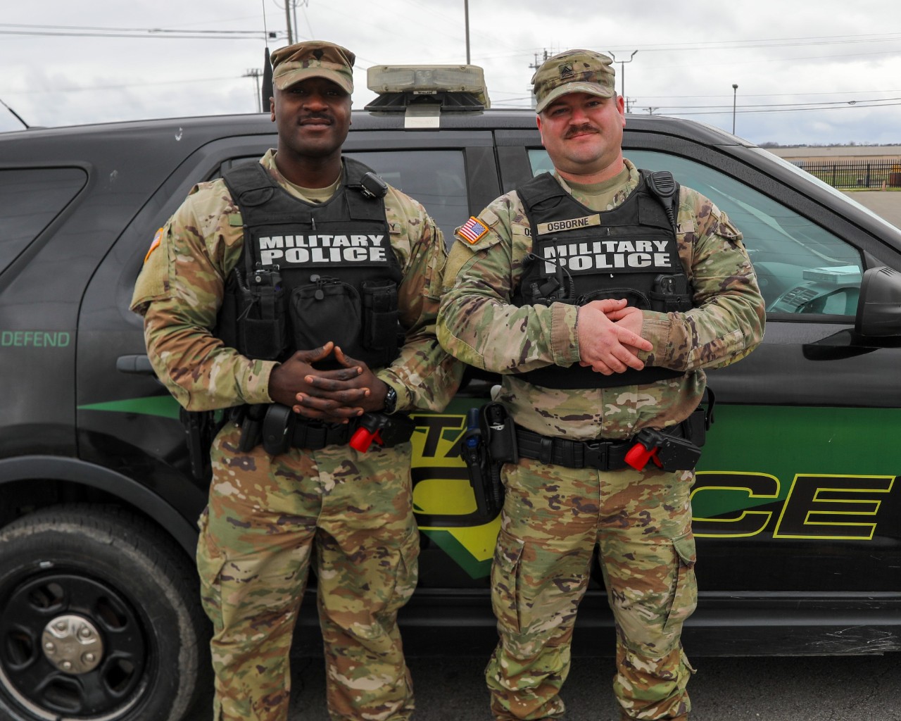 Photo of two military police Soldiers smiling wearing camouflage uniforms with vests that say military police. The men are standing in front of a military police vehicle.