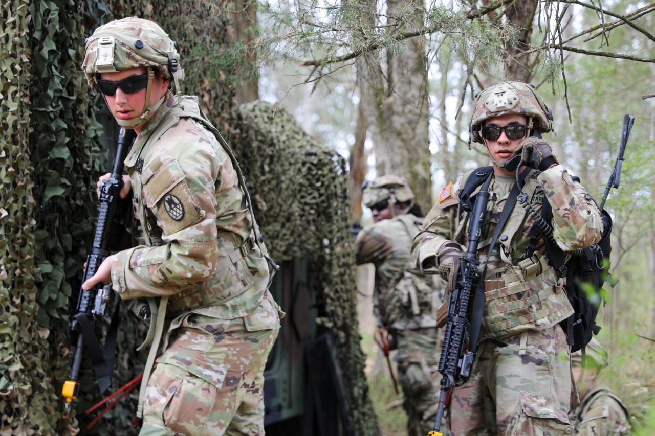 Military Police Soldiers wearing camouflage Army Soldier uniforms patrol through a wooded area. The Soldiers are carrying weapons and one is talking on a radio. 