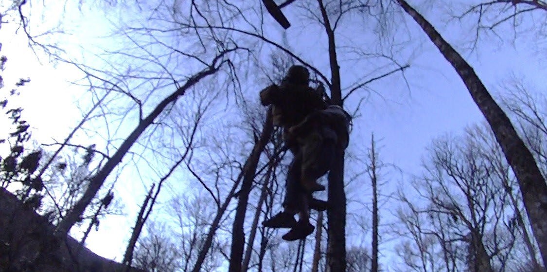 Photo of a wooded area with a silhouette of a Soldier and a hiker being hoisted up through the trees to a helicopter.