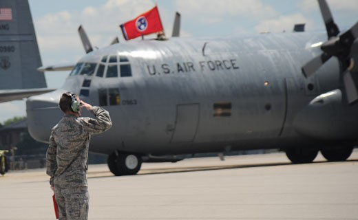 A Tennessee Airman salutes on an aircraft runway as a Tennessee National Guard Aircraft sits in the distance with the State of Tennessee's Flag flies above the aircraft.