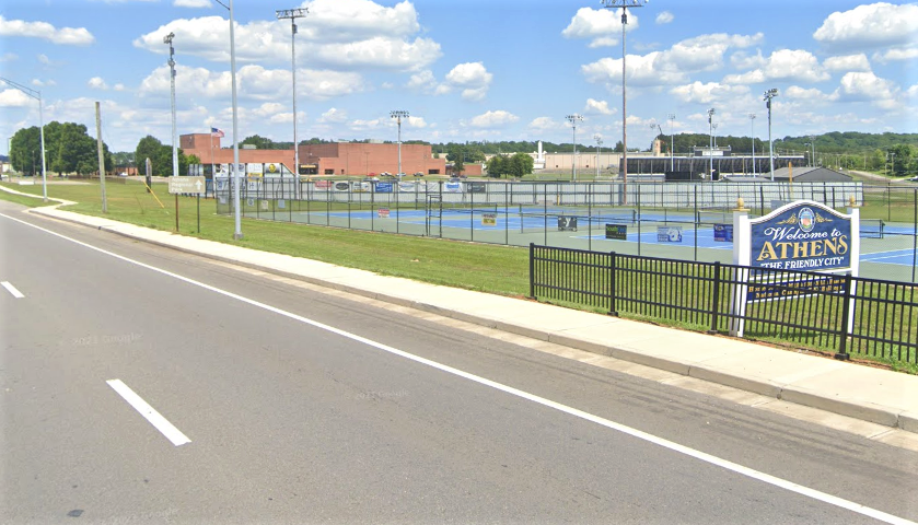 Athens Multimodal Pedestrial Improvements A roadway runs alongside fenced outdoor courts and athletic facilities in a small town setting.