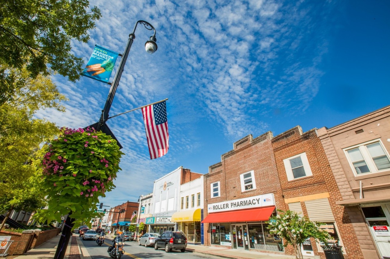 Erwin Downtown Revitalization and Broadband American flags wave above a sunny small-town main street lined with brick storefronts and local shops, as cars and a motorcycle pass beneath a bright blue, cloud-streaked sky.