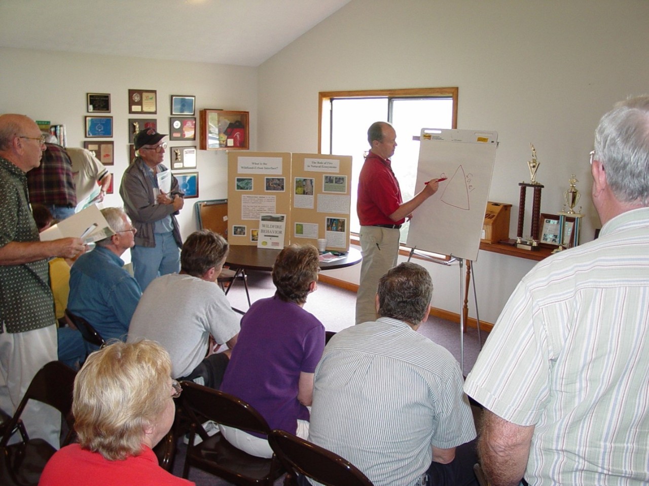Hazard Mitigation and Community Education Grants Man presents to a small group indoors, drawing a diagram on a flip chart while attendees listen and review display boards.