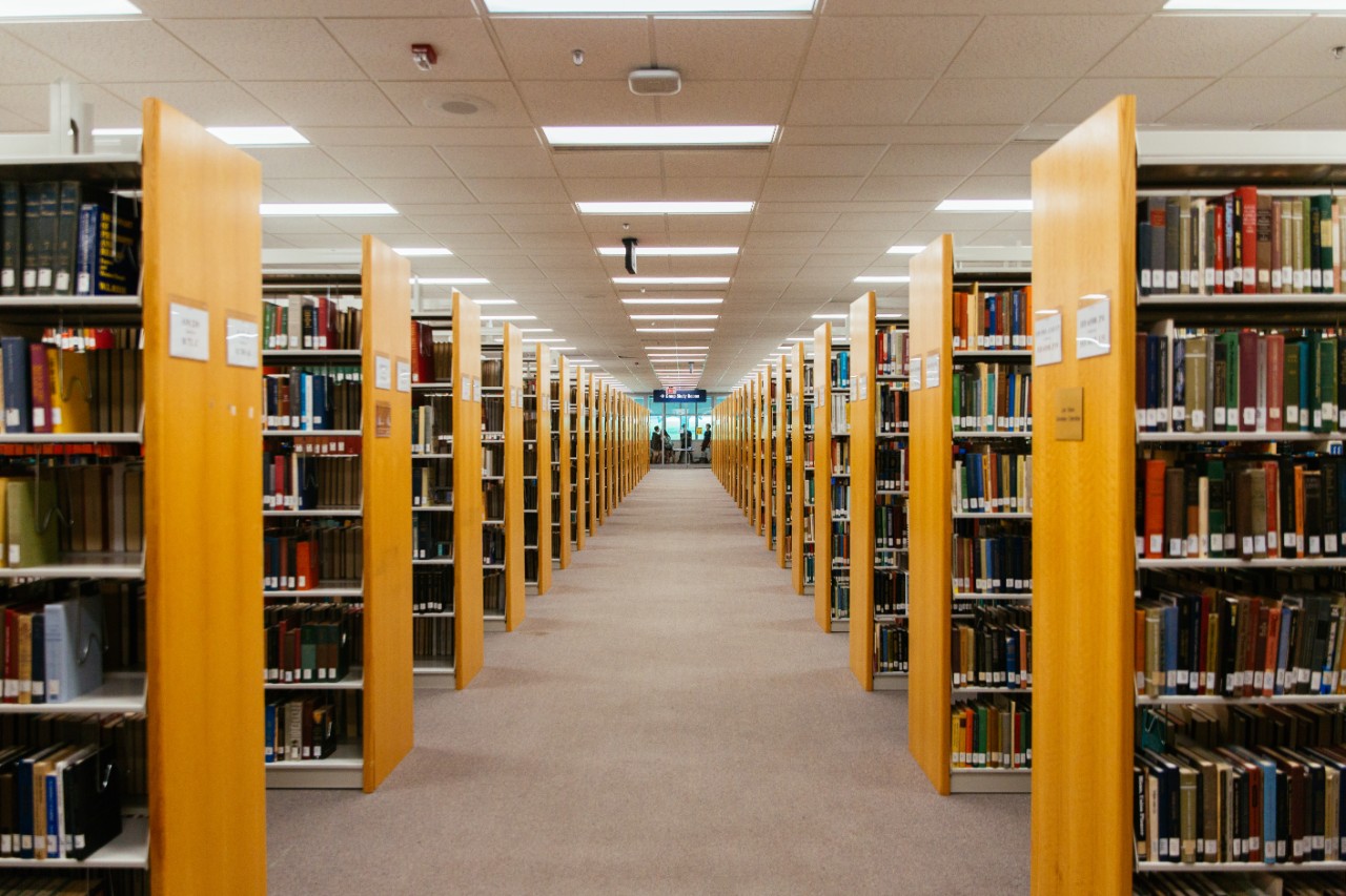 Mildred 2 Rows of bookshelves extend down a long aisle inside a library.