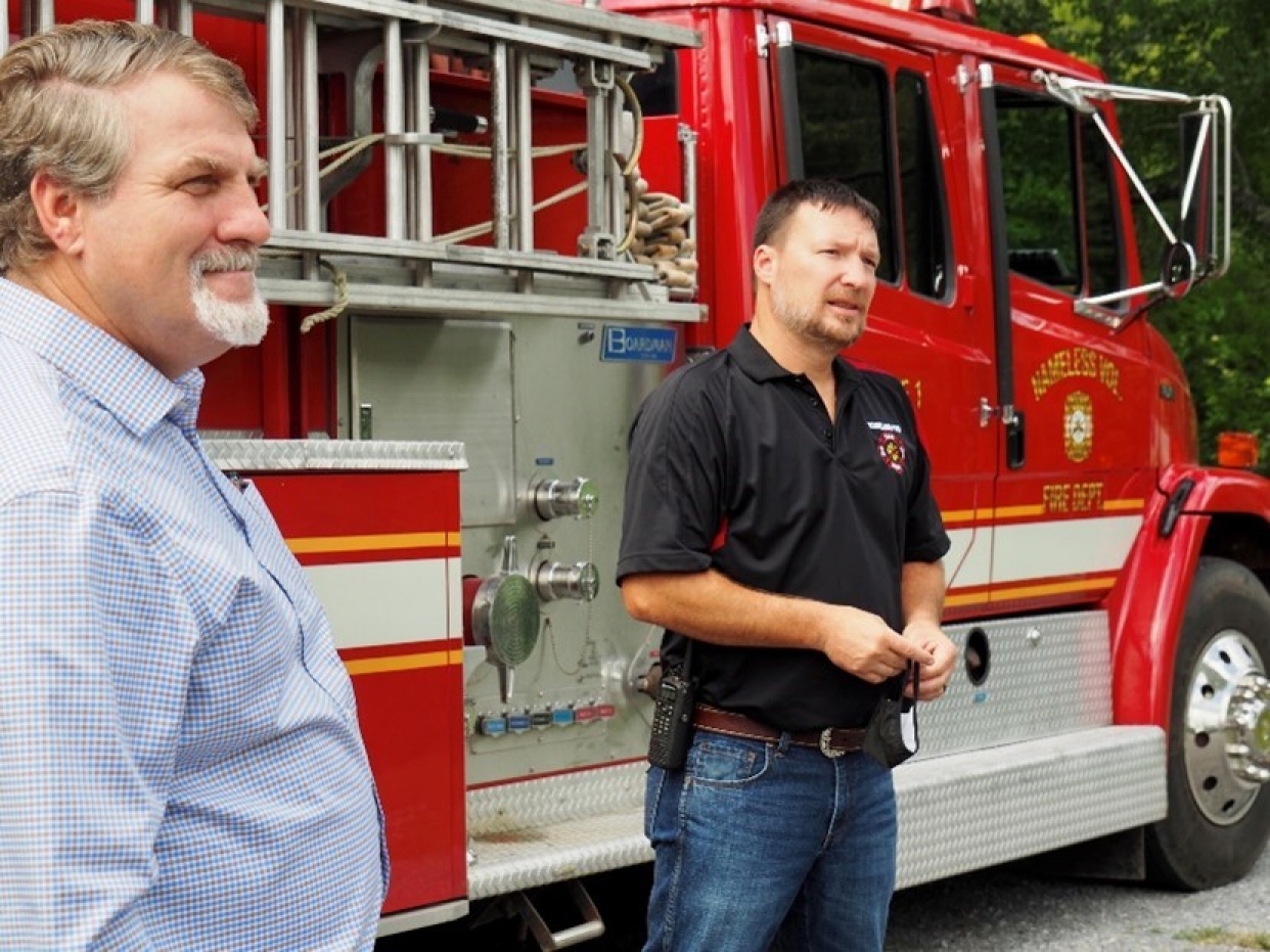 Nameless Volunteer Fire Department PPE Two adults stand beside a fire engine while talking outdoors.
