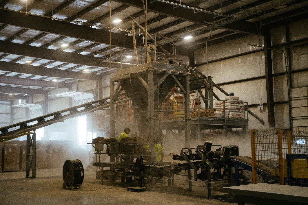 An industrial facility contains large processing equipment, conveyor systems, and workers operating machinery indoors.
