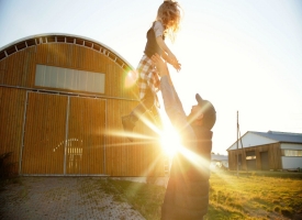 adult man holding young child up in the air, rural barn and sunset behind them