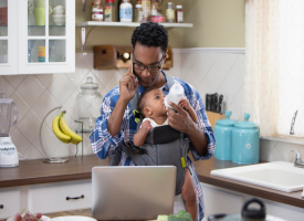 Untitled design - 5 A dad is giving a baby a bottle while on the phone and looking at his laptop